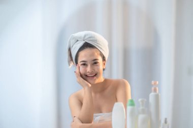 Saçına havlu sarılmış neşeli bir kadın gösteriş için otururken yüz kremi sürüyor. Cilt bakımı ürünleri ve ayna görünür olup, rahatlatıcı ve sıcak ortamlarda kişisel bakım rutinini vurgular.