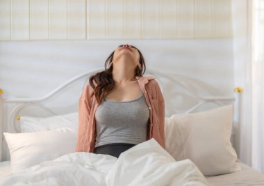 Beyaz bir yatakta otururken kollarını uzatan genç bir kadın rahatlatıcı ve ferahlatıcı bir hava yayıyor. Yumuşak ışıklandırma ve rahat ortam sakin bir sabah atmosferi yaratır..