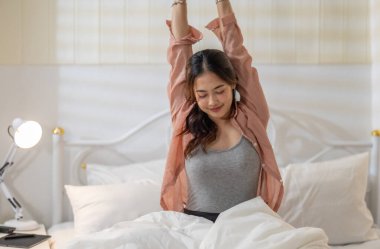 Beyaz bir yatakta otururken kollarını uzatan genç bir kadın rahatlatıcı ve ferahlatıcı bir hava yayıyor. Yumuşak ışıklandırma ve rahat ortam sakin bir sabah atmosferi yaratır..