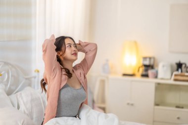 Beyaz bir yatakta otururken kollarını uzatan genç bir kadın rahatlatıcı ve ferahlatıcı bir hava yayıyor. Yumuşak ışıklandırma ve rahat ortam sakin bir sabah atmosferi yaratır..