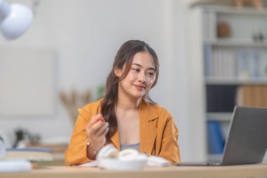 Günümüz ev ofisinin düzenli masasında dizüstü bilgisayar üzerinde çalışan sarı ceketli genç bir kadın. Görüntü üretkenliği, odaklanmayı ve ev ortamında rahat bir çalışma sunar..