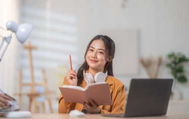 Günümüz ev ofisinin düzenli masasında dizüstü bilgisayar üzerinde çalışan sarı ceketli genç bir kadın. Görüntü üretkenliği, odaklanmayı ve ev ortamında rahat bir çalışma sunar..