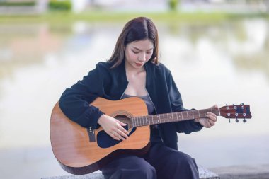 Genç bir kadın halka açık alanda akustik gitar çalıyor, müziğe dalmış. Serin açık hava ortamı sakin ve sanatsal atmosfere katkı sağlayarak yaratıcı enerji ve performans için mükemmel bir zemin oluşturur..