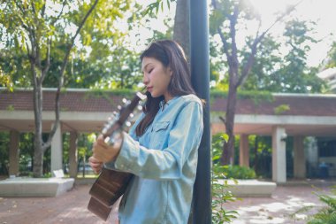 Genç bir kadın halka açık alanda akustik gitar çalıyor, müziğe dalmış. Serin açık hava ortamı sakin ve sanatsal atmosfere katkı sağlayarak yaratıcı enerji ve performans için mükemmel bir zemin oluşturur..