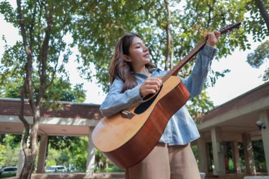 Genç bir kadın halka açık alanda akustik gitar çalıyor, müziğe dalmış. Serin açık hava ortamı sakin ve sanatsal atmosfere katkı sağlayarak yaratıcı enerji ve performans için mükemmel bir zemin oluşturur..