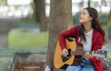 Genç bir kadın halka açık alanda akustik gitar çalıyor, müziğe dalmış. Serin açık hava ortamı sakin ve sanatsal atmosfere katkı sağlayarak yaratıcı enerji ve performans için mükemmel bir zemin oluşturur..