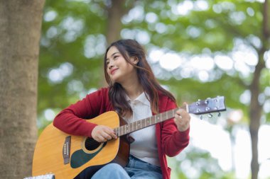 Genç bir kadın halka açık alanda akustik gitar çalıyor, müziğe dalmış. Serin açık hava ortamı sakin ve sanatsal atmosfere katkı sağlayarak yaratıcı enerji ve performans için mükemmel bir zemin oluşturur..