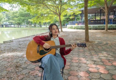 Genç bir kadın halka açık alanda akustik gitar çalıyor, müziğe dalmış. Serin açık hava ortamı sakin ve sanatsal atmosfere katkı sağlayarak yaratıcı enerji ve performans için mükemmel bir zemin oluşturur..