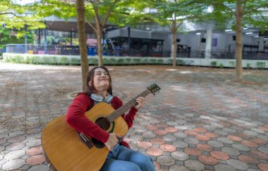 Genç bir kadın halka açık alanda akustik gitar çalıyor, müziğe dalmış. Serin açık hava ortamı sakin ve sanatsal atmosfere katkı sağlayarak yaratıcı enerji ve performans için mükemmel bir zemin oluşturur..