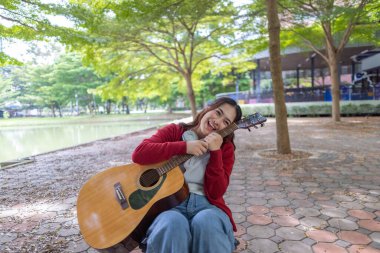 Genç bir kadın halka açık alanda akustik gitar çalıyor, müziğe dalmış. Serin açık hava ortamı sakin ve sanatsal atmosfere katkı sağlayarak yaratıcı enerji ve performans için mükemmel bir zemin oluşturur..