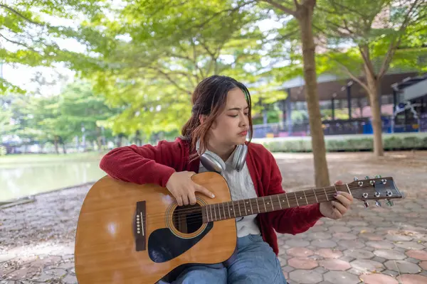 Genç bir kadın halka açık alanda akustik gitar çalıyor, müziğe dalmış. Serin açık hava ortamı sakin ve sanatsal atmosfere katkı sağlayarak yaratıcı enerji ve performans için mükemmel bir zemin oluşturur..