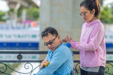 Bir kadın, tekerlekli sandalyede oturan bir adama, kaslarındaki gerilimi azaltmak için masaj topu kullanarak terapi amaçlı masaj yapar. Huzurlu park kurulumu bakım, destek ve refah temalarının altını çiziyor.