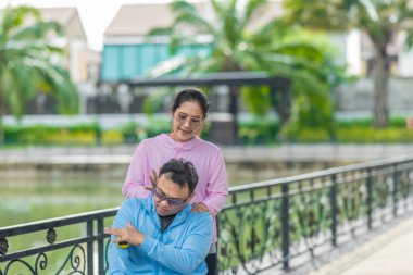 Bir kadın, tekerlekli sandalyede oturan bir adama, kaslarındaki gerilimi azaltmak için masaj topu kullanarak terapi amaçlı masaj yapar. Huzurlu park kurulumu bakım, destek ve refah temalarının altını çiziyor.
