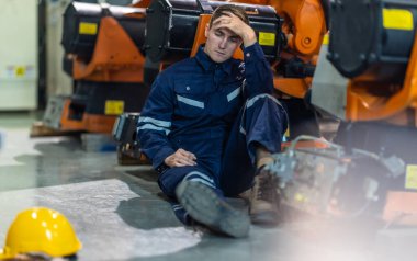 Fabrika teknisyeni sanayi ortamında yerde oturuyor, bitkin ve bunalmış görünüyor. İşçi alnına kadar el ile makinelere yaslanır, yorgunluk ve iş stresi taşır.