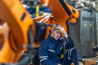 Fabrika teknisyeni sanayi ortamında yerde oturuyor, bitkin ve bunalmış görünüyor. İşçi alnına kadar el ile makinelere yaslanır, yorgunluk ve iş stresi taşır.