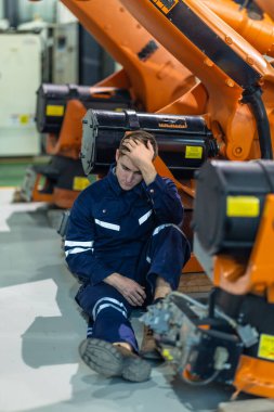 Fabrika teknisyeni sanayi ortamında yerde oturuyor, bitkin ve bunalmış görünüyor. İşçi alnına kadar el ile makinelere yaslanır, yorgunluk ve iş stresi taşır.