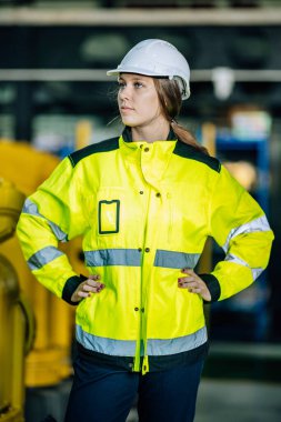 Parlak yeşil ceketli ve beyaz şapkalı bir işçi endüstriyel bir ortamda dikkatle duruyor. Ayar iyi ışıklandırılmış, gösterişli makineler ve güvenlik ekipmanları var..