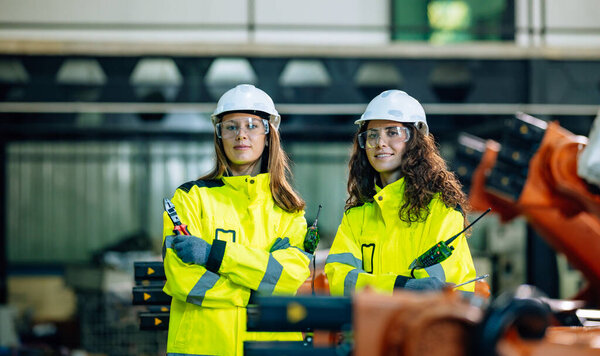 Two women engineers stand confidently in bright yellow safety jackets and helmets at a manufacturing plant, overseeing robotic machinery while ensuring safety and efficiency.