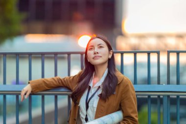 Kasketli ve kahverengi ceketli kadın mimar, geleceğe emin bir şekilde bakarken mimari planlar tutuyor. Modern kentsel zemin üzerine kurulu yenilikçi düşük karbonlu binaların tasarımında rol