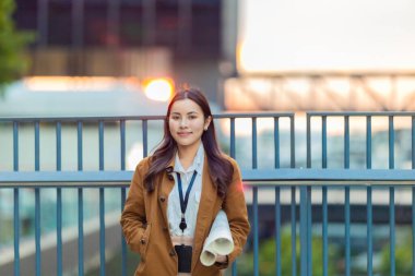 Kasketli ve kahverengi ceketli kadın mimar, geleceğe emin bir şekilde bakarken mimari planlar tutuyor. Modern kentsel zemin üzerine kurulu yenilikçi düşük karbonlu binaların tasarımında rol