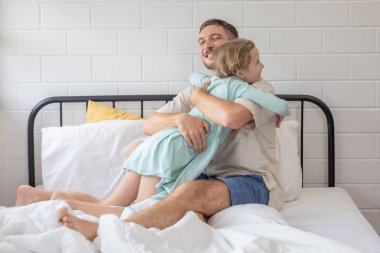Bir adam ve kızı beyaz çarşaflarla kaplı bir yatakta kucaklaşıyorlar. Oda aydınlık ve her ikisi de gülümseyerek huzurlu bir sabahta sıcak bir aile ortamı yaratıyor..
