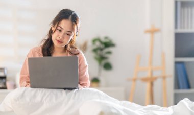 A woman sits comfortably in her cozy bedroom, focused on her laptop while seated on her bed. Soft lighting enhances the peaceful ambiance as she enjoys her evening routine. clipart