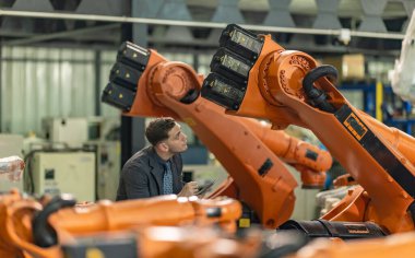 Fabrika sahibi bir üretim tesisinde endüstriyel robotik kolun kalite denetimini yapıyor. Sahnede modern otomasyon teknolojisi ve sanayi operasyonlarında hassas mühendislik sergileniyor..