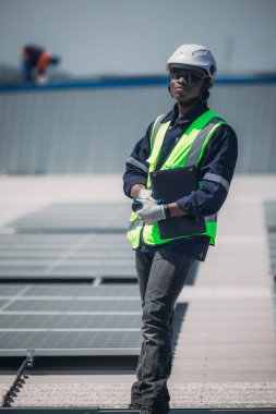Güvenlik kaskı ve yansıtıcı yelek giyen bir işçi çatıda duruyor ve güneş panellerini inceliyor. Gün aydınlık güneş ışığıyla aydınlık. Güneş tesisini vurguluyor..