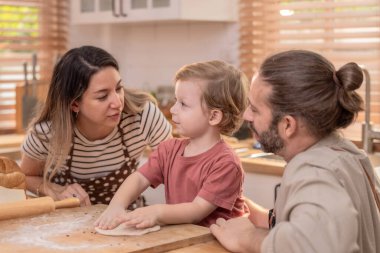 A family is happily engaged in baking at home, with a child playfully participating as they prepare dough on a wooden surface surrounded by bread. clipart