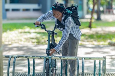 Genç siyah bir kadın bisikletini rafa kilitliyor, çevre dostu ve sürdürülebilir şehir ulaşımını gösteriyor. Sahne, sorumluluk sahibi ve çevreye duyarlı bir işe gidip gelme seçeneğini vurguluyor..