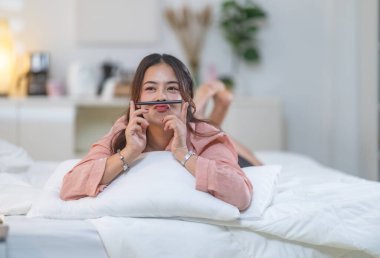 Yumuşak ışıklı bir yatak odasındaki beyaz bir yatakta rahat bir şekilde yatan genç bir kadın. Sahne sakin ve rahat bir atmosfer sunuyor, yaşam tarzı, rahatlık ve rahatlık temaları için mükemmel..