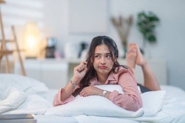 Yumuşak ışıklı bir yatak odasındaki beyaz bir yatakta rahat bir şekilde yatan genç bir kadın. Sahne sakin ve rahat bir atmosfer sunuyor, yaşam tarzı, rahatlık ve rahatlık temaları için mükemmel..