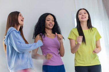 Üç kültürlü arkadaştan oluşan canlı bir grup içeride dans seansının keyfini çıkarıyor. Neşeli ve rahat görünüyorlar, günlük atletik kıyafetler giyiyorlar, arkadaşlığı, eğlenceyi ve hareketi vurguluyorlar.