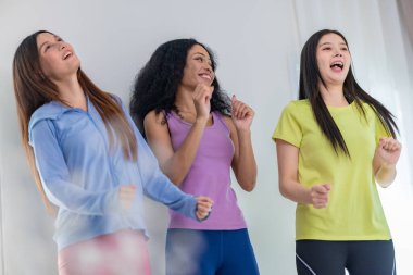 Üç kültürlü arkadaştan oluşan canlı bir grup içeride dans seansının keyfini çıkarıyor. Neşeli ve rahat görünüyorlar, günlük atletik kıyafetler giyiyorlar, arkadaşlığı, eğlenceyi ve hareketi vurguluyorlar.