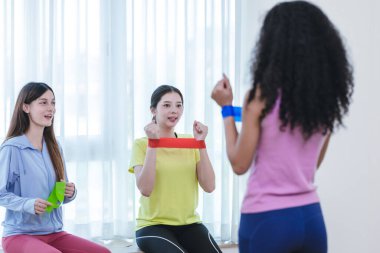 Bir grup çok kültürlü kadın, kapalı bir ortamda birlikte direniş bandı egzersizi yapıyorlar. Görüntü takım çalışmasını, uygunluğu ve destekleyici bir ortamda egzersiz yapmanın zevkini yakalar..