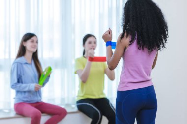 Bir grup çok kültürlü kadın, kapalı bir ortamda birlikte direniş bandı egzersizi yapıyorlar. Görüntü takım çalışmasını, uygunluğu ve destekleyici bir ortamda egzersiz yapmanın zevkini yakalar..