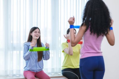 Bir grup çok kültürlü kadın, kapalı bir ortamda birlikte direniş bandı egzersizi yapıyorlar. Görüntü takım çalışmasını, uygunluğu ve destekleyici bir ortamda egzersiz yapmanın zevkini yakalar..