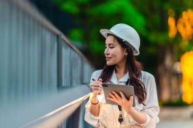 Beyaz güvenlik kaskı ve iş kıyafeti giyen Asyalı kadın mimar yeşil şehir ticari projelerini denetlerken elinde bir tablet tutuyor. Modern, sürdürülebilir ticari kalkınmayı vurgula