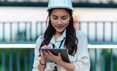 Beyaz güvenlik kaskı ve iş kıyafeti giyen Asyalı kadın mimar yeşil şehir ticari projelerini denetlerken elinde bir tablet tutuyor. Modern, sürdürülebilir ticari kalkınmayı vurgula