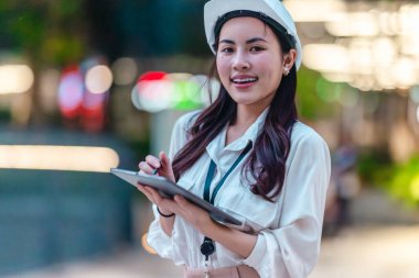 Beyaz güvenlik kaskı ve iş kıyafeti giyen Asyalı kadın mimar yeşil şehir ticari projelerini denetlerken elinde bir tablet tutuyor. Modern, sürdürülebilir ticari kalkınmayı vurgula