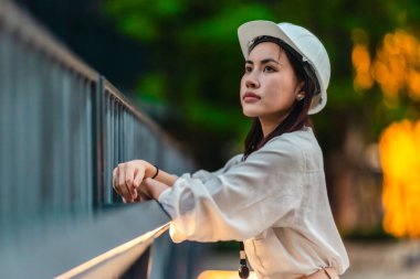 Beyaz güvenlik kaskı ve iş kıyafeti giyen Asyalı kadın mimar yeşil şehir ticari projelerini denetlerken elinde bir tablet tutuyor. Modern, sürdürülebilir ticari kalkınmayı vurgula