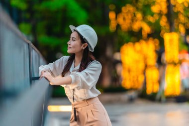 Beyaz güvenlik kaskı ve iş kıyafeti giyen Asyalı kadın mimar yeşil şehir ticari projelerini denetlerken elinde bir tablet tutuyor. Modern, sürdürülebilir ticari kalkınmayı vurgula