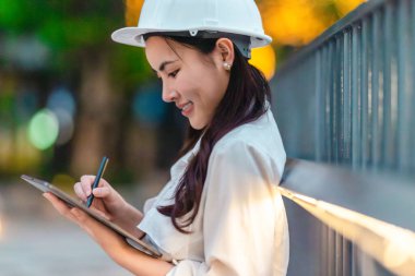 Beyaz güvenlik kaskı ve iş kıyafeti giyen Asyalı kadın mimar yeşil şehir ticari projelerini denetlerken elinde bir tablet tutuyor. Modern, sürdürülebilir ticari kalkınmayı vurgula