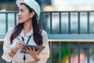 Beyaz güvenlik kaskı ve iş kıyafeti giyen Asyalı kadın mimar yeşil şehir ticari projelerini denetlerken elinde bir tablet tutuyor. Modern, sürdürülebilir ticari kalkınmayı vurgula