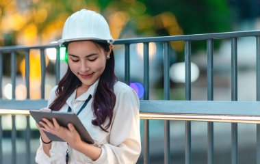 Beyaz güvenlik kaskı ve iş kıyafeti giyen Asyalı kadın mimar yeşil şehir ticari projelerini denetlerken elinde bir tablet tutuyor. Modern, sürdürülebilir ticari kalkınmayı vurgula