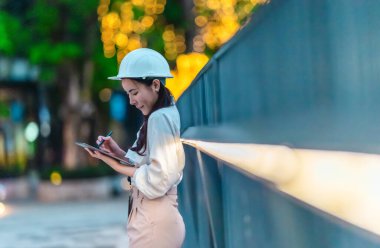Beyaz güvenlik kaskı ve iş kıyafeti giyen Asyalı kadın mimar yeşil şehir ticari projelerini denetlerken elinde bir tablet tutuyor. Modern, sürdürülebilir ticari kalkınmayı vurgula