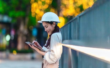 Beyaz güvenlik kaskı ve iş kıyafeti giyen Asyalı kadın mimar yeşil şehir ticari projelerini denetlerken elinde bir tablet tutuyor. Modern, sürdürülebilir ticari kalkınmayı vurgula