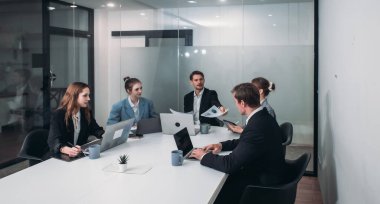 İş adamları konferans odasında toplandı. Resmi giysiler içindeki ekip üyeleri, belgeleri tartışıyor ve dizüstü bilgisayar kullanıyor, işbirlikçi ve üretken çalışma ortamı yaratıyorlar..