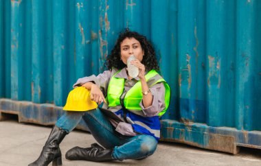 Yorgun bir kadın işçi, elinde su şişesi ve sert bir şapka tutarken alnından ter silerken nakliye konteynırının karşısında oturur. Yorgunluk, işyeri stresi, fiziksel işçiliğin etkisi
