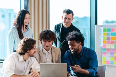 Çeşitli başlangıç takımları bilgisayar ekranına gülümseyen video konferansına katılıyor. Parlak çalışma alanı, takım çalışması, yenilik ve işbirliğini yansıtan renkli yapışkan notlarla dolu bir beyaz tahtaya sahip.